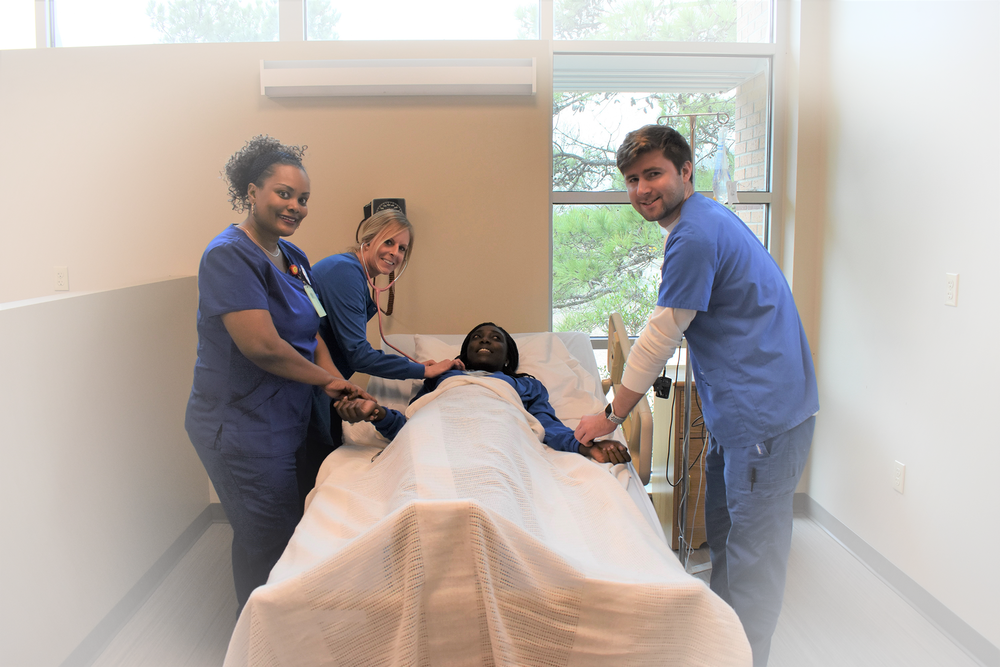 Registered Nurse- A female nurse in blue scrubs holding a blue clipboard with her co-workers standing behind her smiling